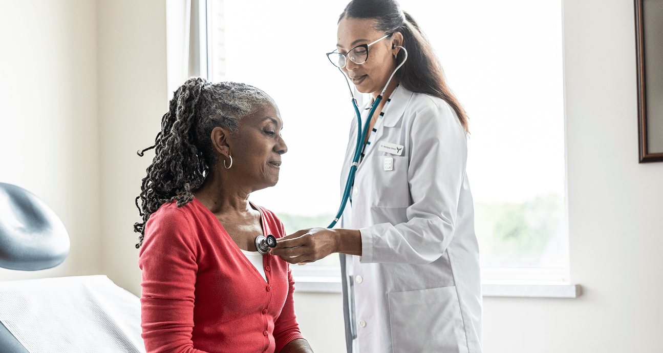A healthcare professional in a white lab coat uses a stethoscope to examine an older adult patient wearing a red cardigan while the patient sits on an exam table in a bright medical examination room.