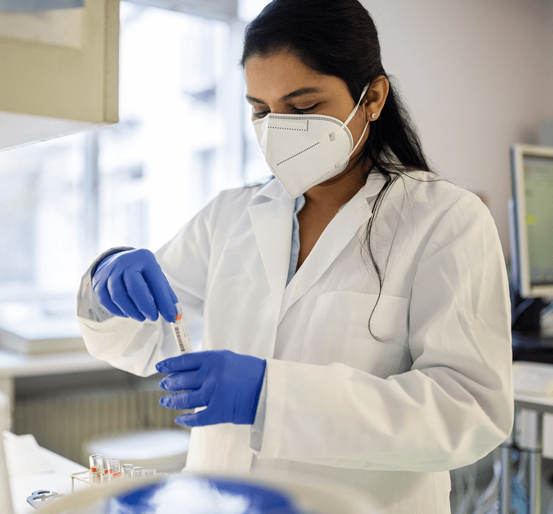 A laboratory researcher wearing a white lab coat, protective mask, and blue gloves carefully handles a test tube in a laboratory.