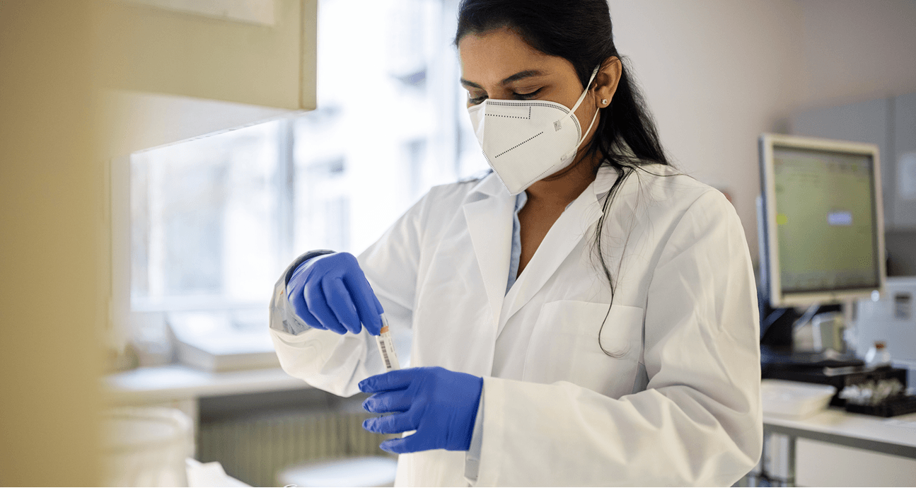 A laboratory researcher wearing a white lab coat, protective mask, and blue gloves carefully handles a test tube in a laboratory.